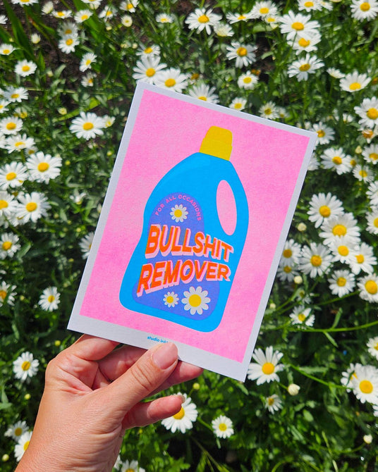 Roze Riso art print "Bullshit Remover" met blauwe wasmiddelfles, gehouden tegen madeliefjes.