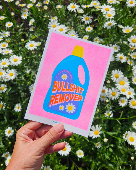 Roze Riso art print "Bullshit Remover" met blauwe wasmiddelfles, gehouden tegen madeliefjes.