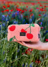 Roze teddy etui met rode kersenprint, zwart label met gouden tekst en rode rits, vastgehouden in een bloemenveld.