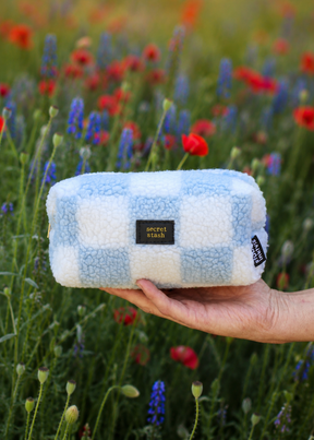 Blauw-wit geruit teddy tasje Secret Stash met gouden letters, vastgehouden in een bloemenveld