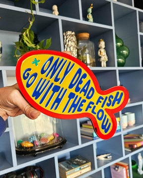 Visvormige gele spiegel wanddecoratie met rode rand en blauwe tekst "Only dead fish go with the flow".