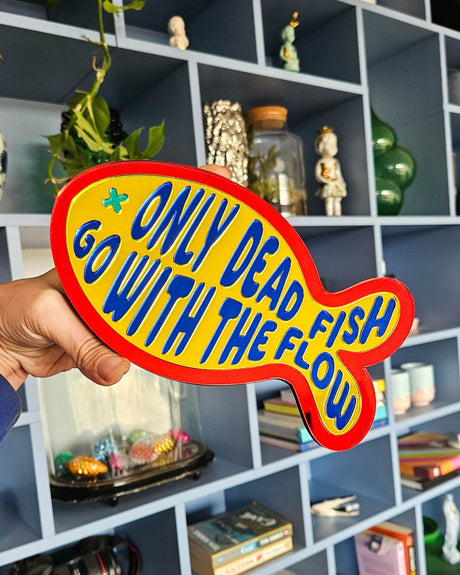 Visvormige gele spiegel wanddecoratie met rode rand en blauwe tekst "Only dead fish go with the flow".
