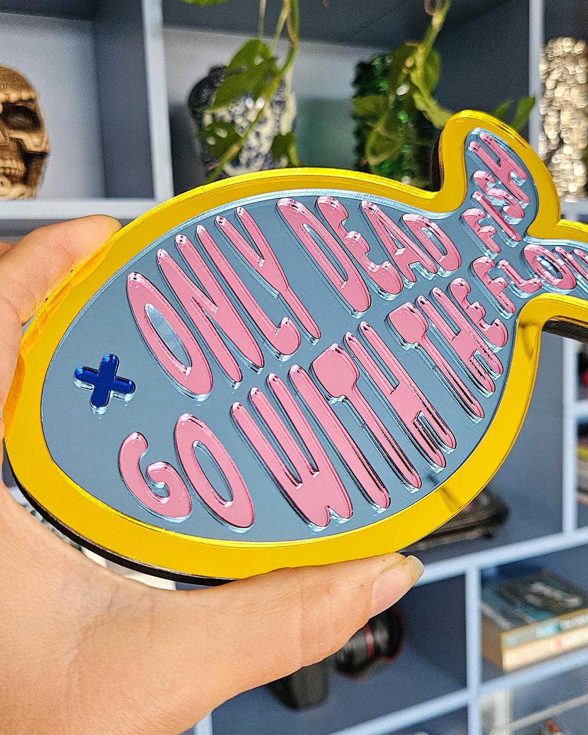 Kleurrijke visvormige spiegel van acryl met "Only dead fish go with the flow" tekst in roze op blauw, gele rand.